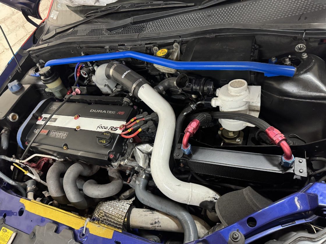 A blue performance engine bar installed across the front of a mk1 Ford Focus engine bay, shown with a black Duratec engine cover, white intake pipe, and various other engine components.